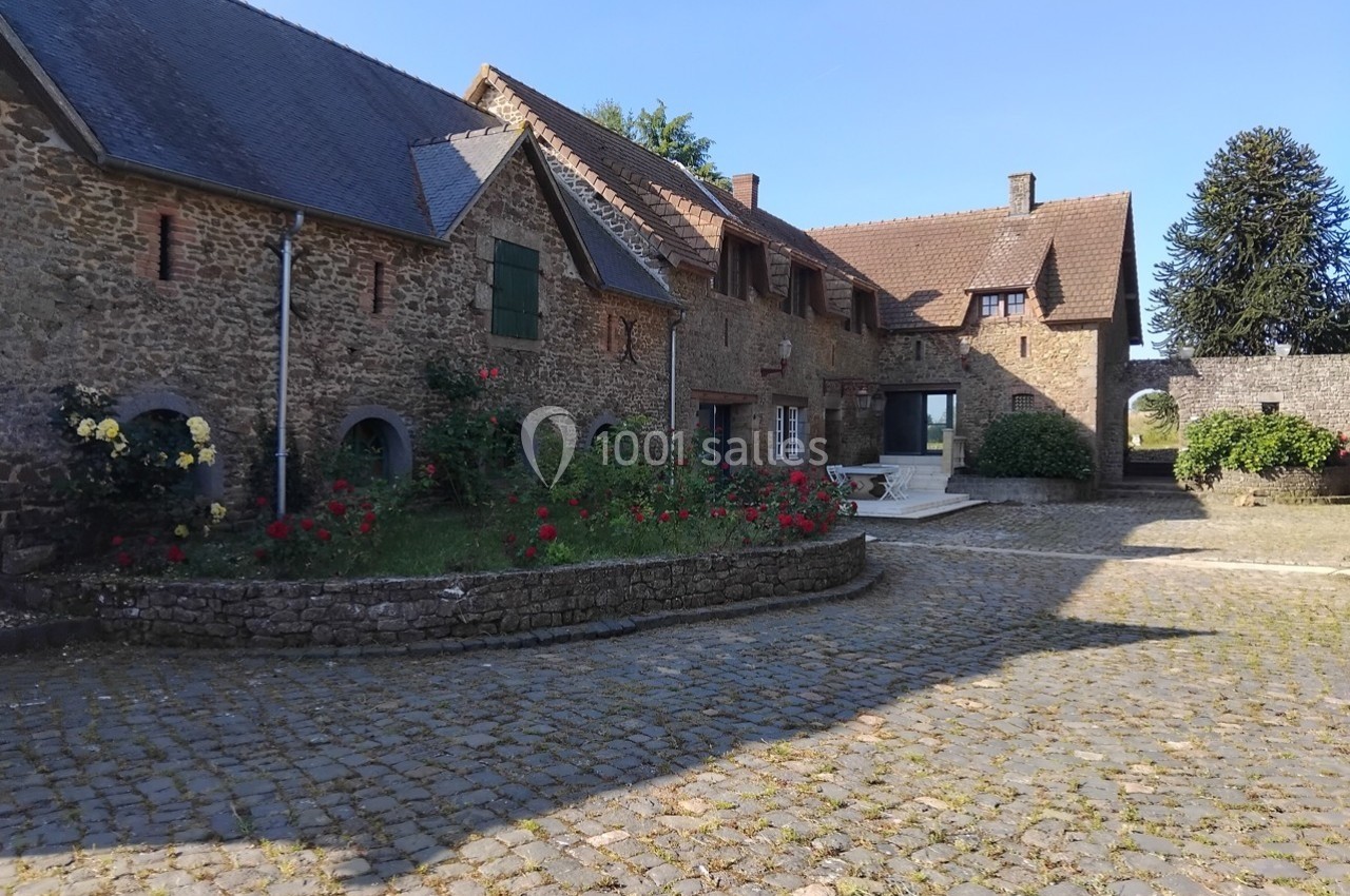 Cour pavée avec bâtiments en pierre, toits en ardoise et jardinières fleuries sous un ciel dégagé.