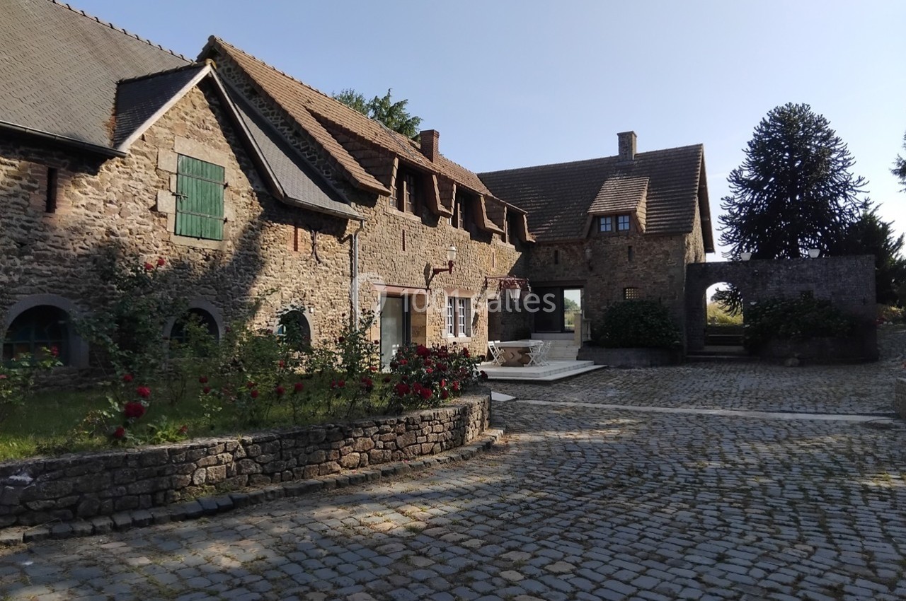 Cour pavée devant un bâtiment en pierre avec des toits en pente, des roses et un arbre en arrière-plan.
