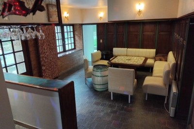 Bâtiment en pierre avec toit en ardoise, petite terrasse en bois, tables blanches et arbres en arrière-plan.