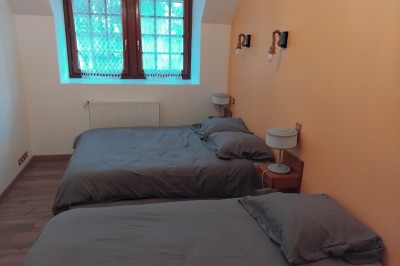 Bâtiment en pierre avec toit en ardoise, petite terrasse en bois, tables blanches et arbres en arrière-plan.