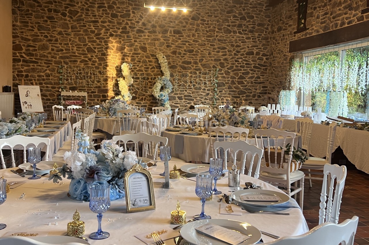 Salle de réception aux murs en pierre apparente, tables rondes nappées de blanc et de bleu dressées pour un banquet, chaises blanches et compositions florales bleues et blanches.