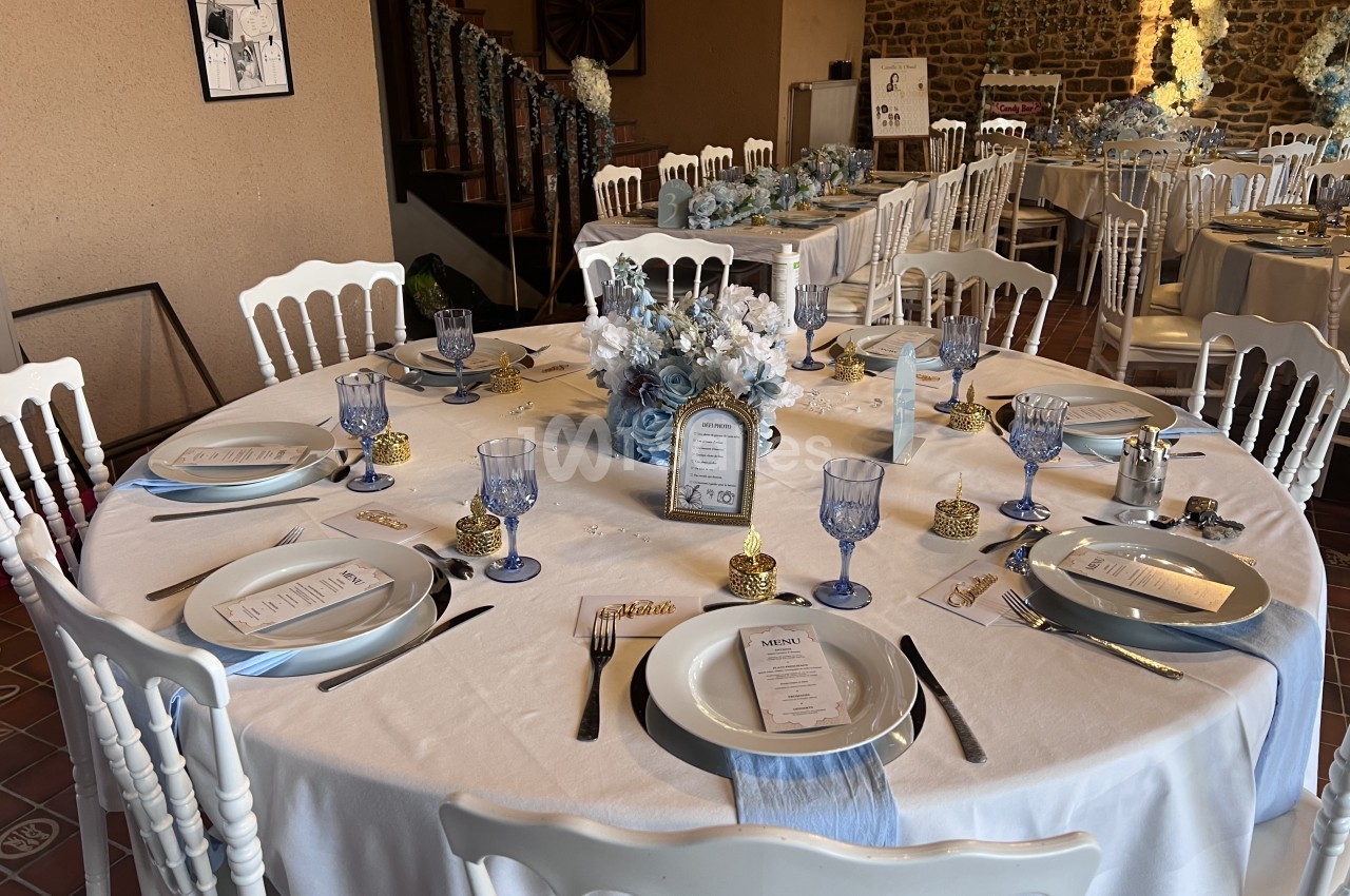Table ronde dressée de nappes blanches, couverts dorés, verres bleus et bouquet de fleurs bleues et blanches, avec d'autres tables en arrière-plan dans une salle aux murs de pierre. Let me verify length: roughly 170 characters, too long. Need ≤125. Table
