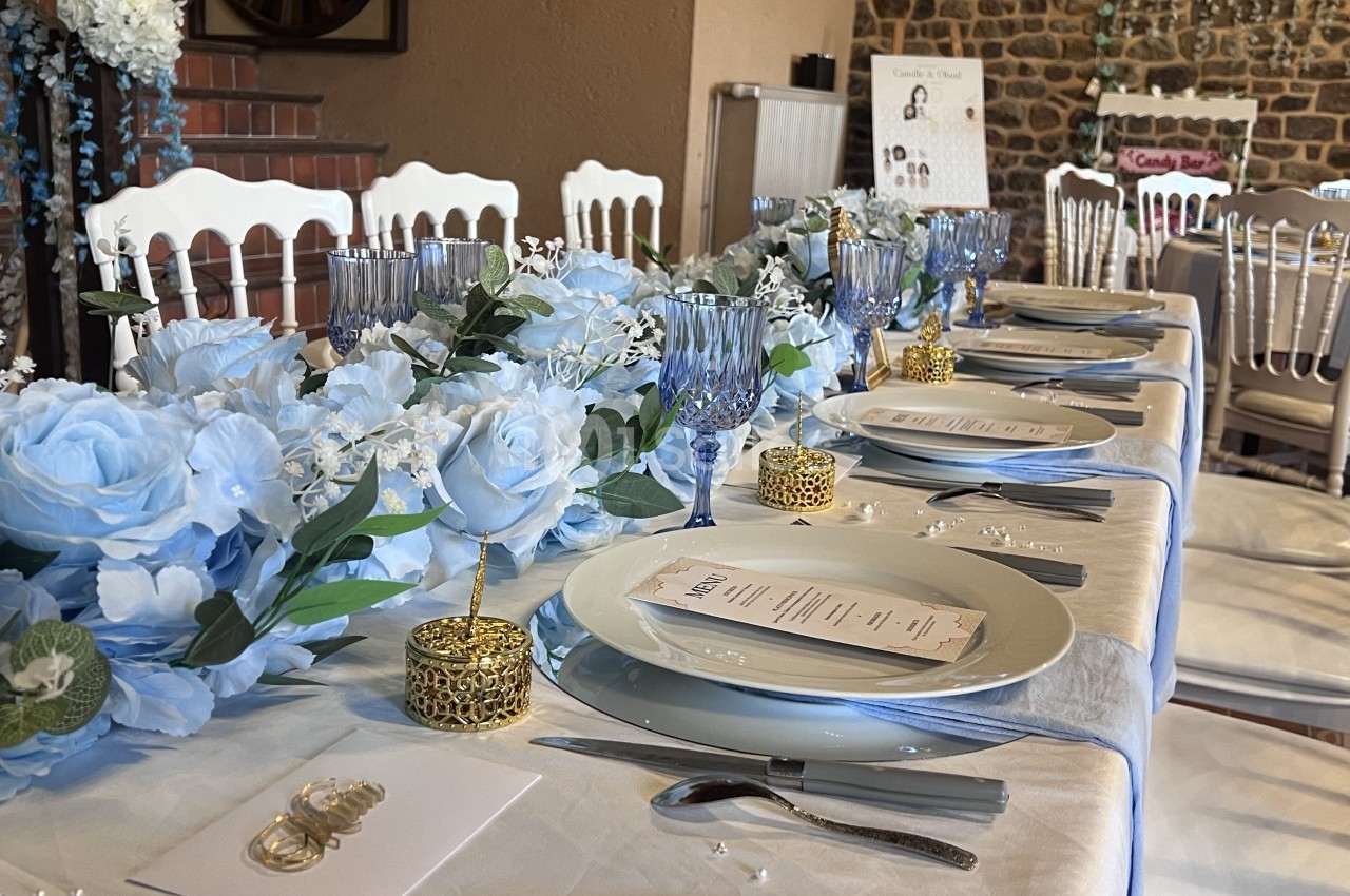 Table de réception dressée de nappes blanches et de bleu pâle, décorée de roses et d'hortensias bleus, de verres à pied bleus et de chaises blanches en bois.