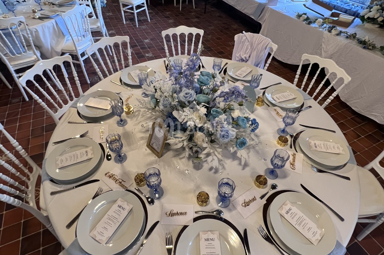 Table ronde dressée de vaisselle blanche, verres bleus et bouquet central de roses et fleurs bleues et blanches, sur sol carrelé en terre cuite.