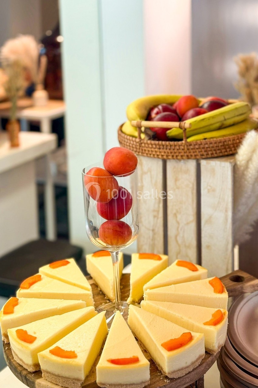 Tranches de cheesecake disposées en cercle sur un plateau, avec des fruits frais en arrière-plan et des prunes dans un verre.