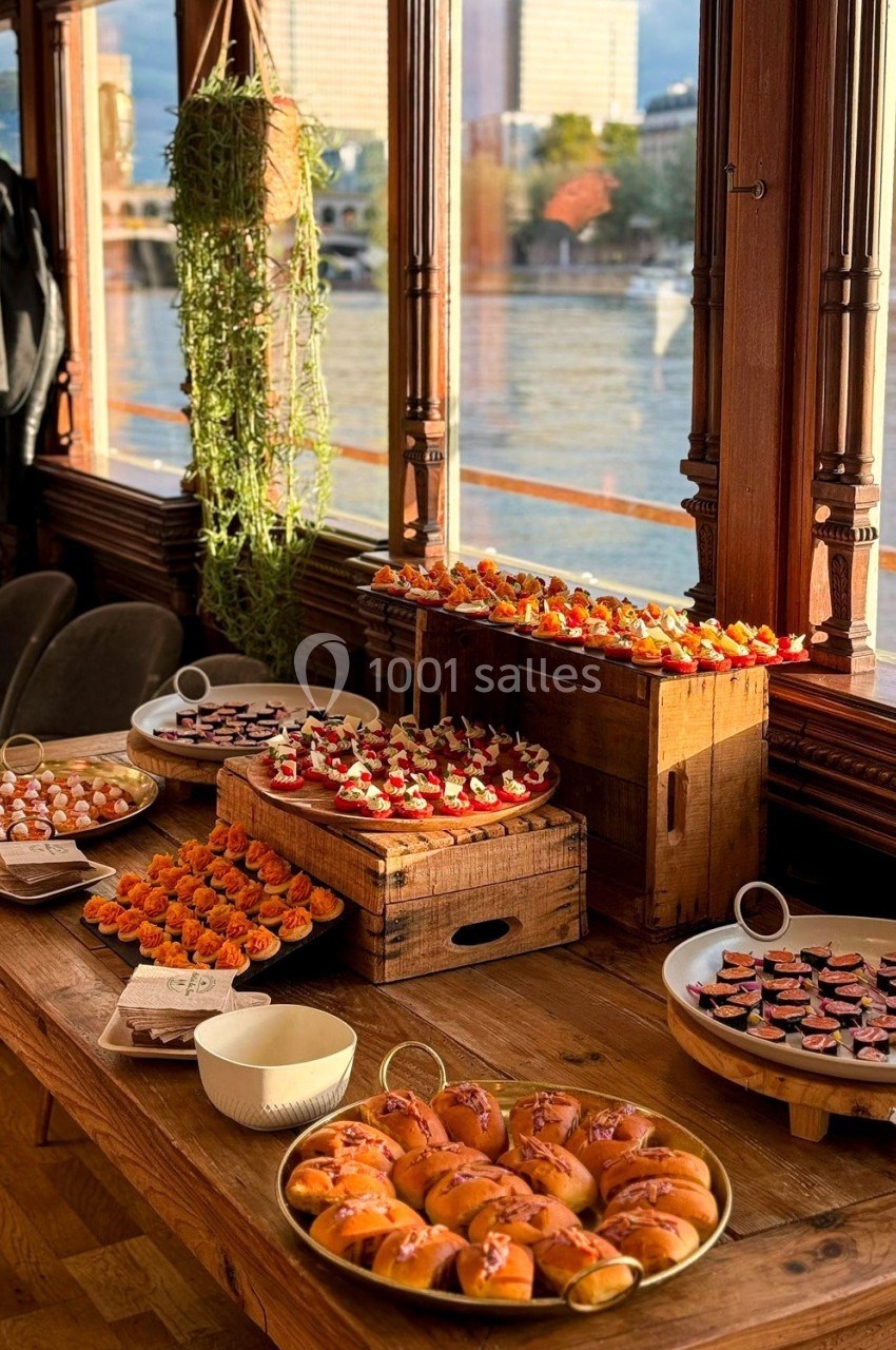 Buffet de canapés variés disposés sur une table en bois près d'une fenêtre avec vue sur un fleuve.