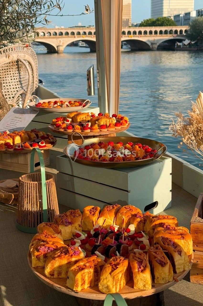 Plateaux de pâtisseries variées disposés sur une table en bois, avec vue sur une rivière et un pont en arrière-plan.
