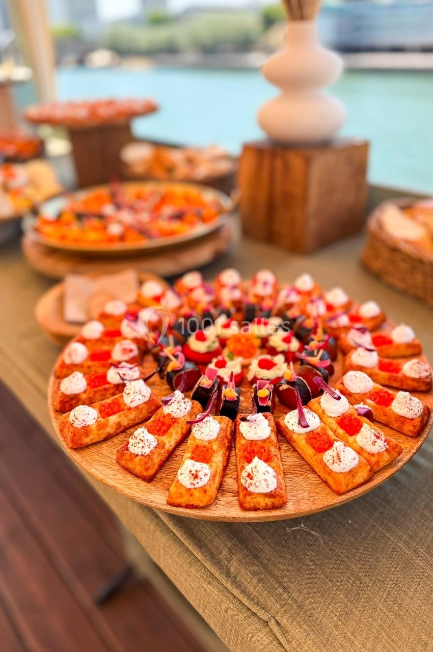 Plateau de toasts variés garnis de fromage, légumes et herbes, présenté sur une table en extérieur près de l'eau.