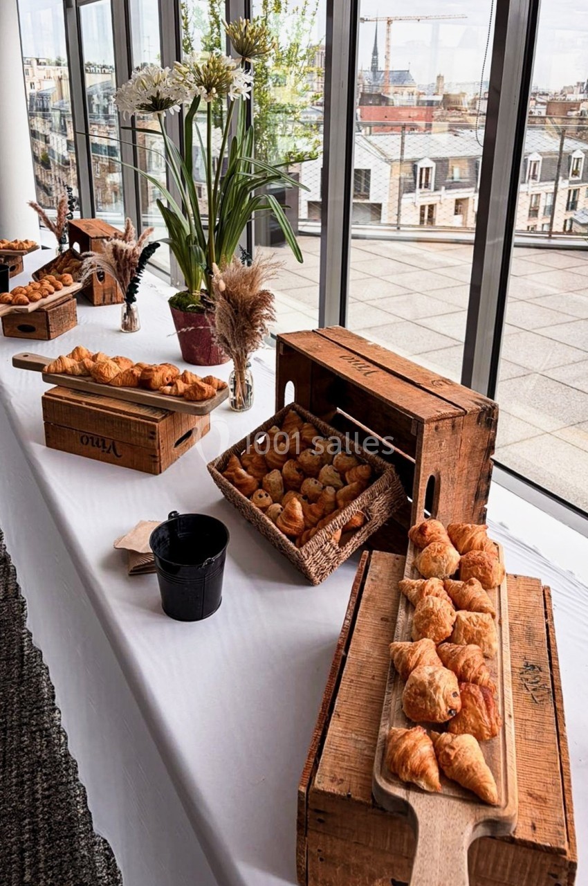 Table dressée avec des viennoiseries sur des planches en bois, entourée de décorations florales, avec vue sur des toits…