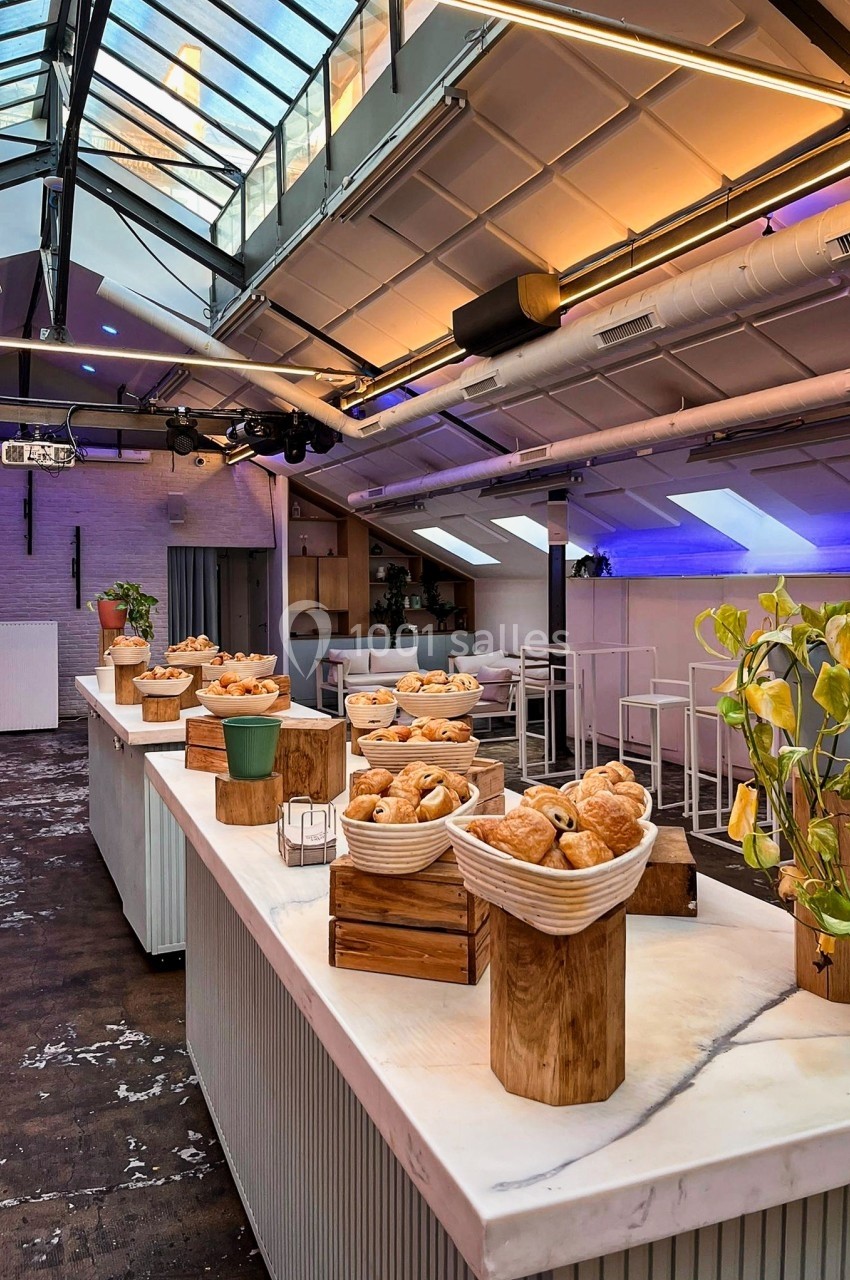 Salle lumineuse avec verrière, tables en marbre garnies de viennoiseries et décorations végétales.