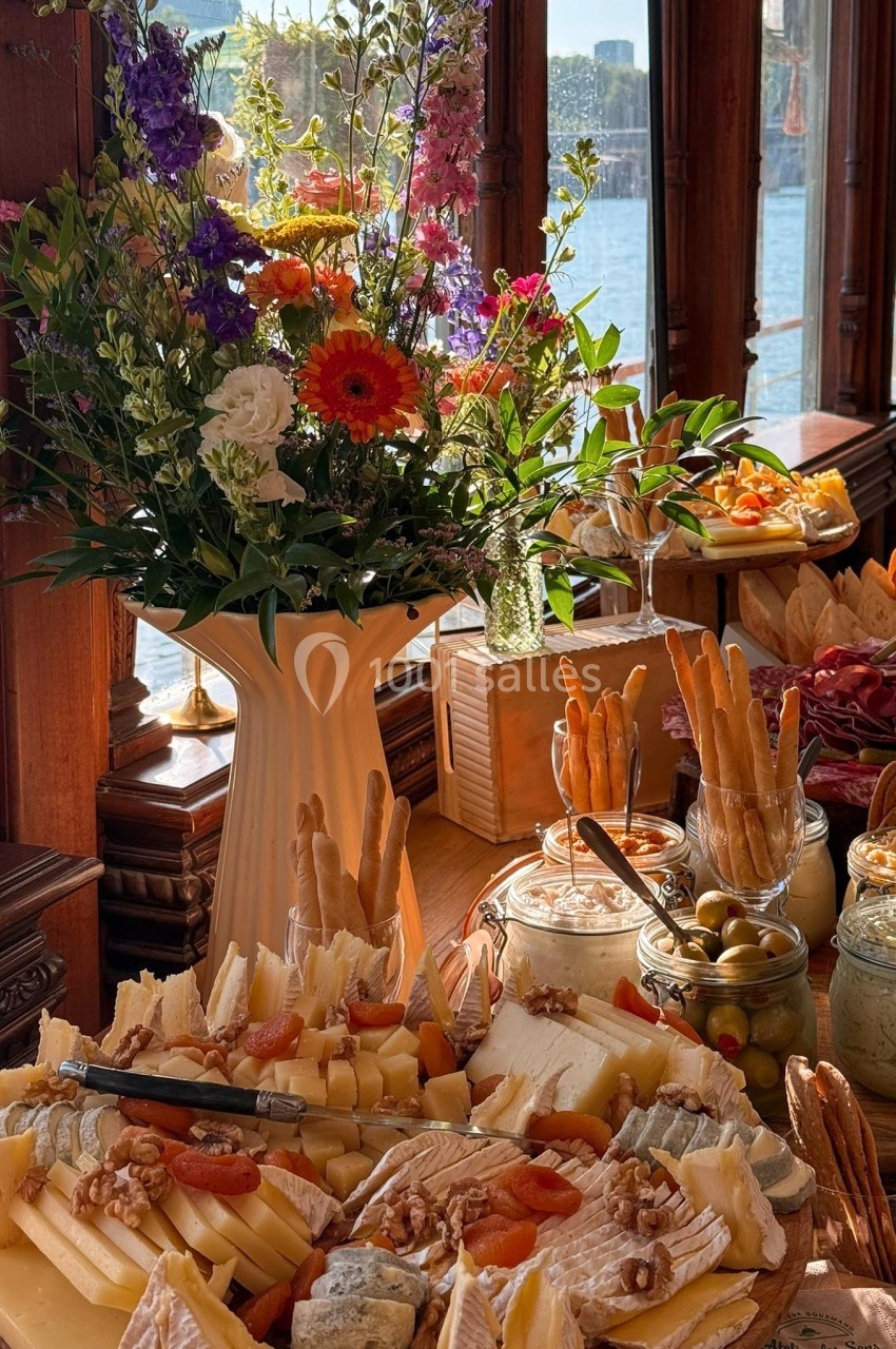 Plateau de fromages variés avec fruits secs, olives et pains, disposé près d'un bouquet de fleurs et d'une fenêtre…