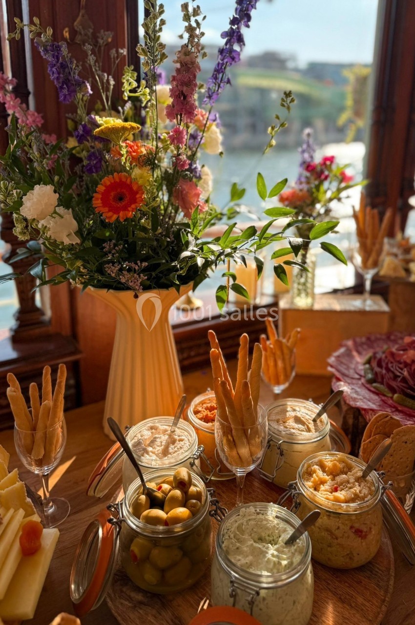 Plateau apéritif avec dips, olives, gressins et fleurs en arrière-plan près d'une fenêtre donnant sur un paysage ensoleillé.