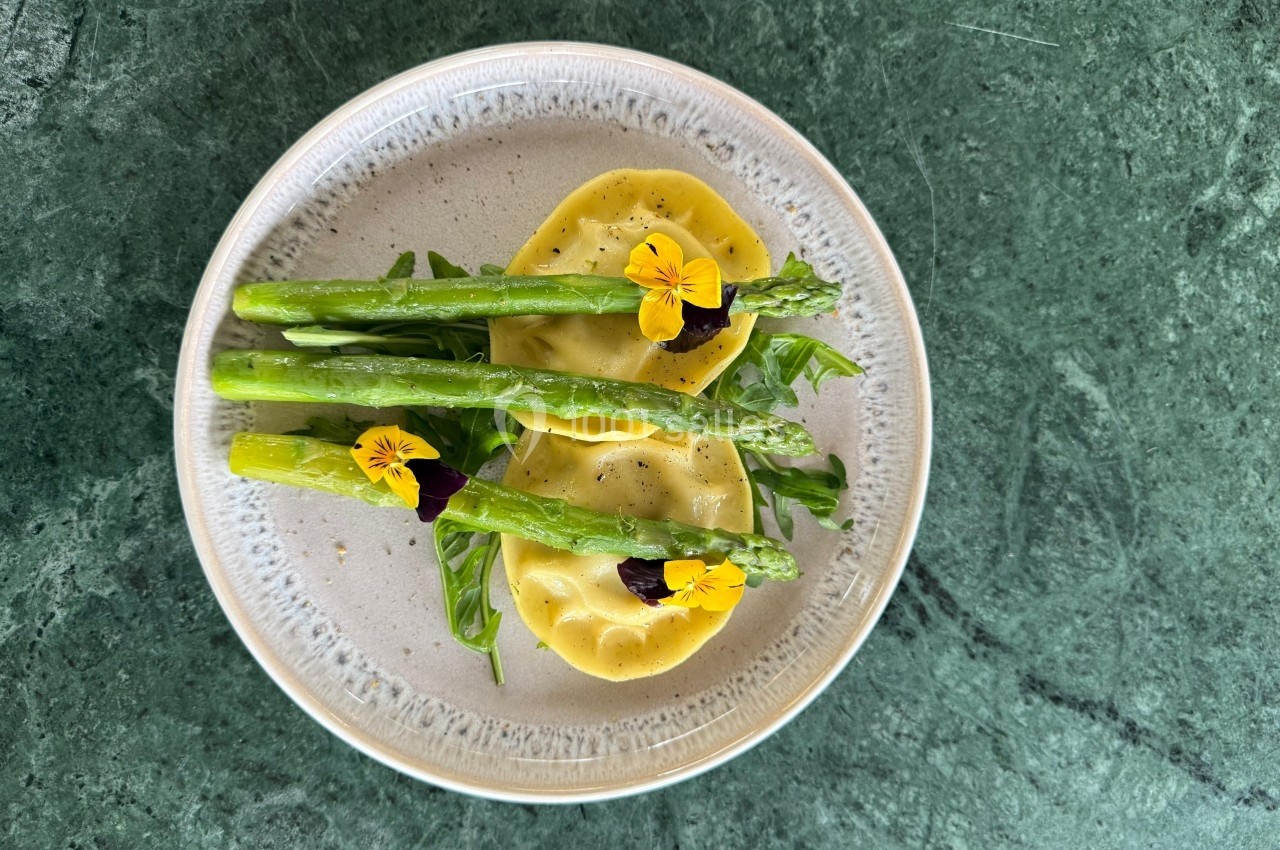 Assiette de raviolis garnis, accompagnés d'asperges, de roquette et de fleurs comestibles sur une table verte.