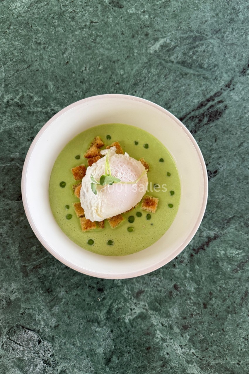 Bol de soupe verte garnie d'un œuf poché, de croûtons et d'une herbe fraîche, posé sur une table en marbre vert.