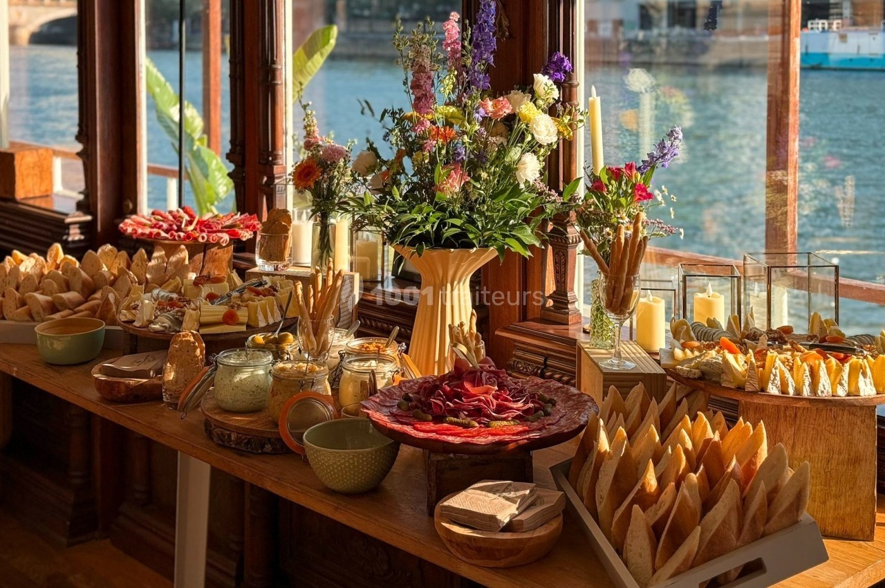 Buffet composé de charcuterie, pains, fromages et fleurs, disposé près de grandes fenêtres avec vue sur l'eau.