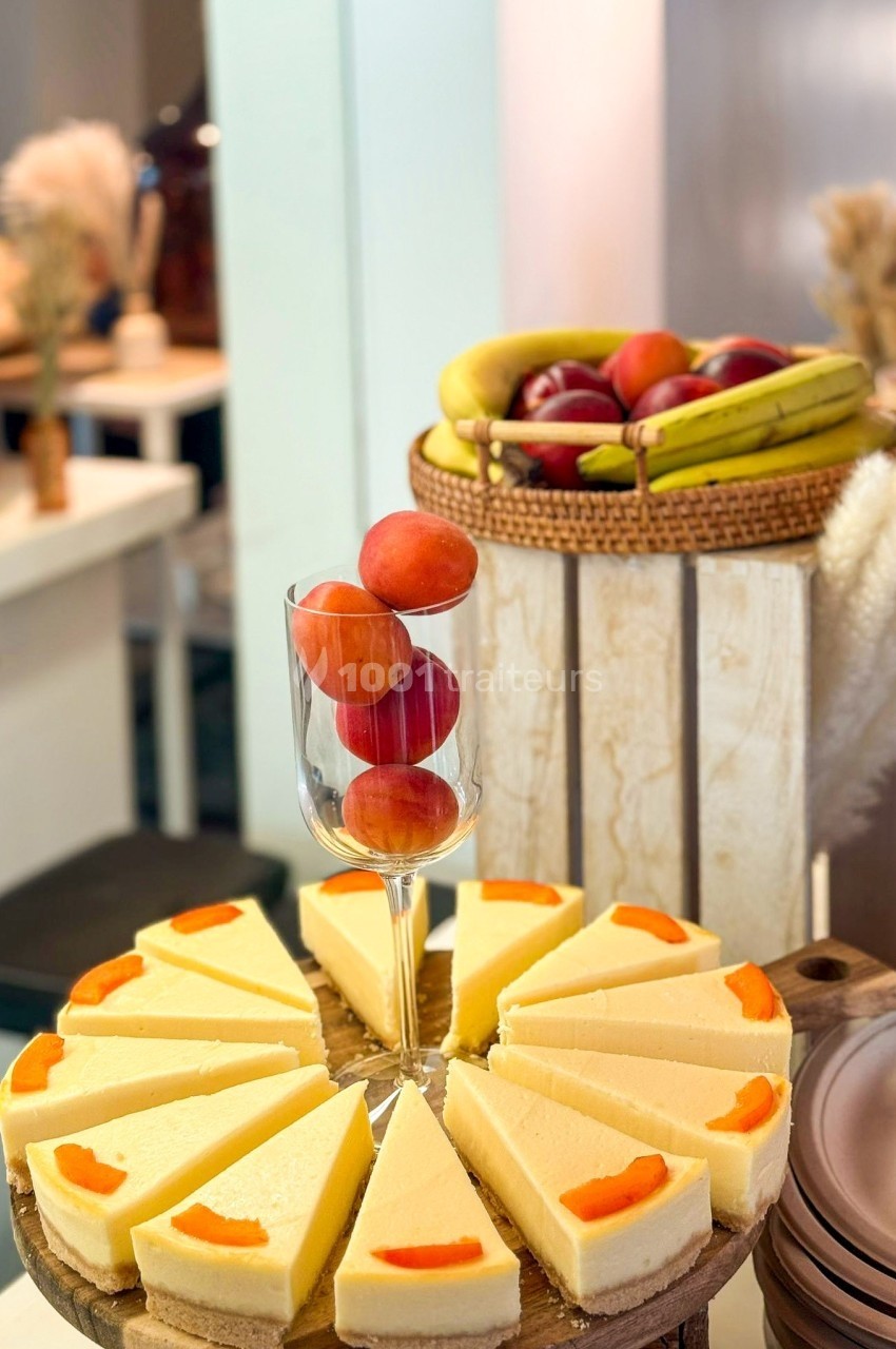 Tranches de cheesecake disposées en cercle, avec des fruits rouges et jaunes dans un verre et un panier en arrière-plan.