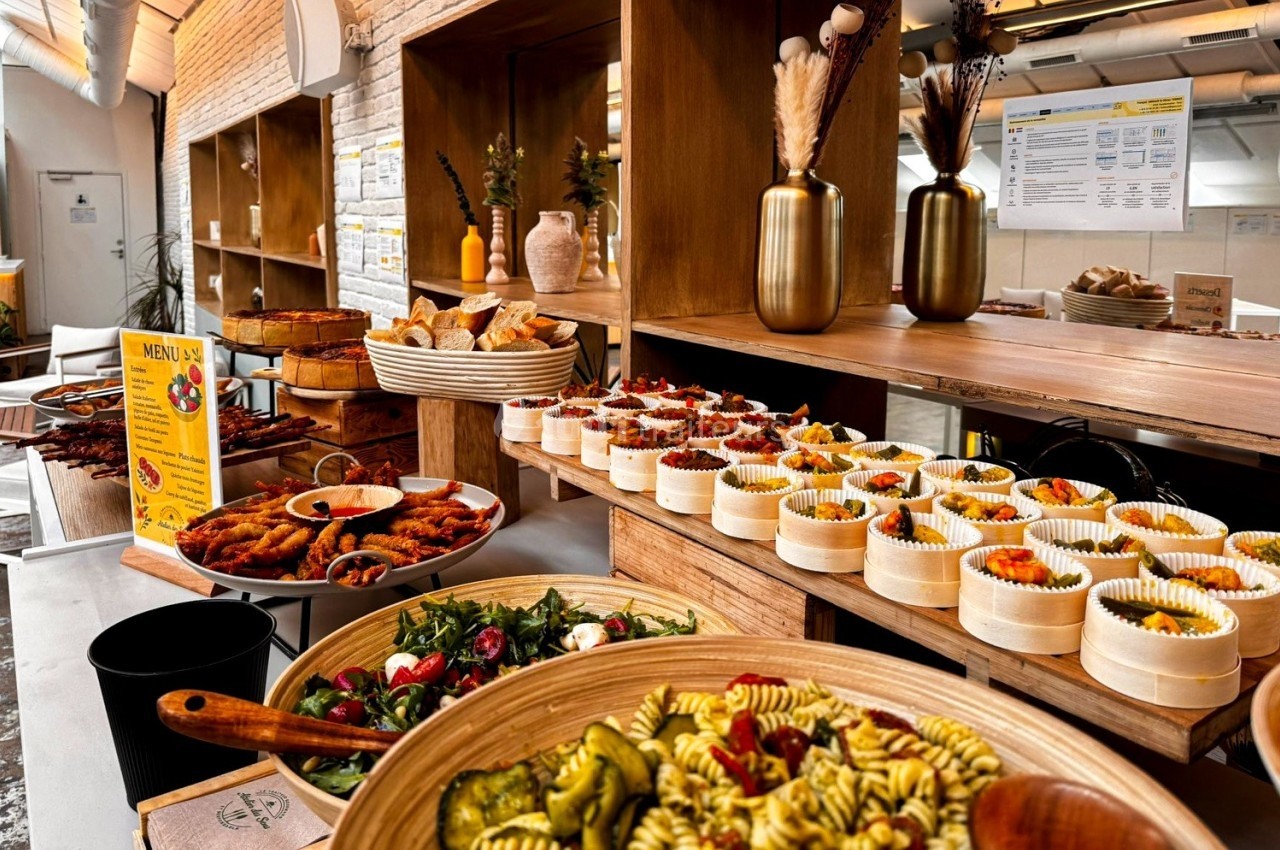 Buffet varié avec salades, plats chauds, pains et desserts présentés sur des étagères en bois dans un espace lumineux.