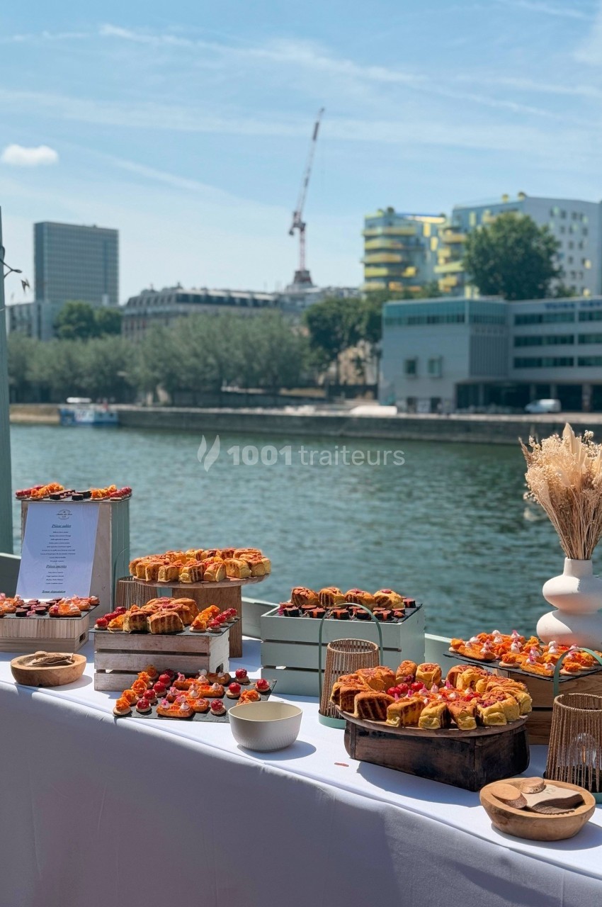 Buffet en plein air avec des amuse-bouches variés disposés sur une table, vue sur une rivière et des bâtiments en arrière…