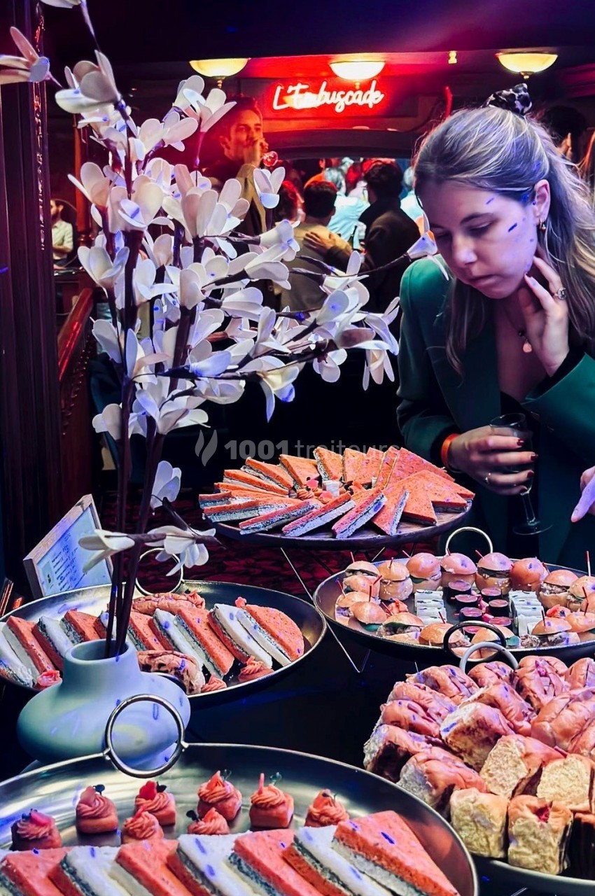 Femme observant un buffet varié de bouchées et sandwiches dans un cadre lumineux avec décoration florale artificielle.