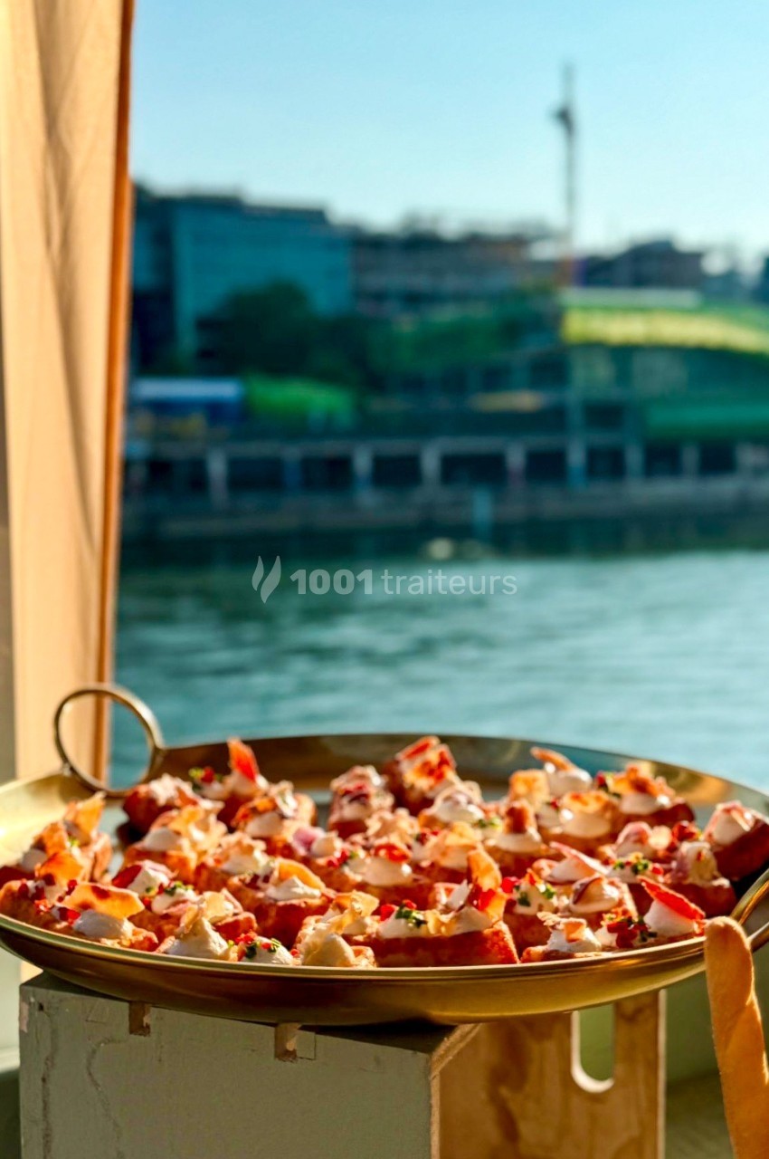 Plat de fruits de mer servi dans une grande poêle, posé près d'une fenêtre avec vue sur une rivière et des bâtiments.