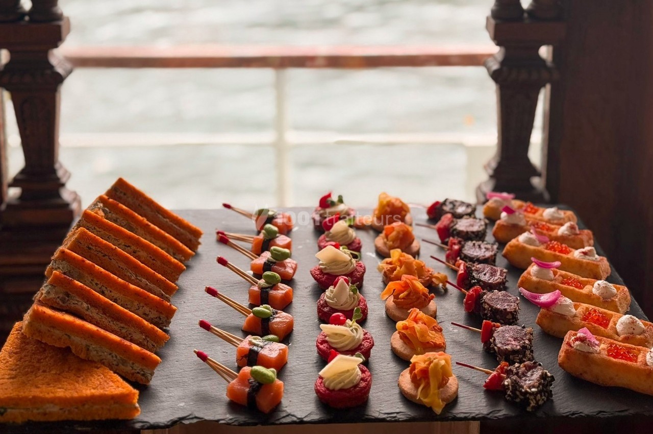 Plateau d'amuse-bouches variés disposés sur une ardoise, avec vue floue sur une fenêtre donnant sur l'extérieur.