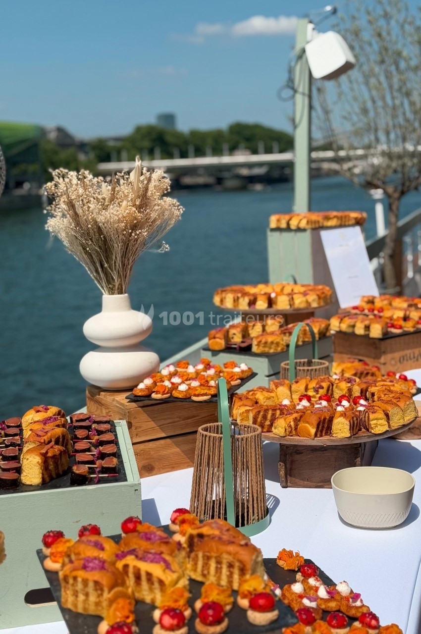Buffet en extérieur avec pâtisseries variées disposées sur des présentoirs en bois, près d'une rivière.