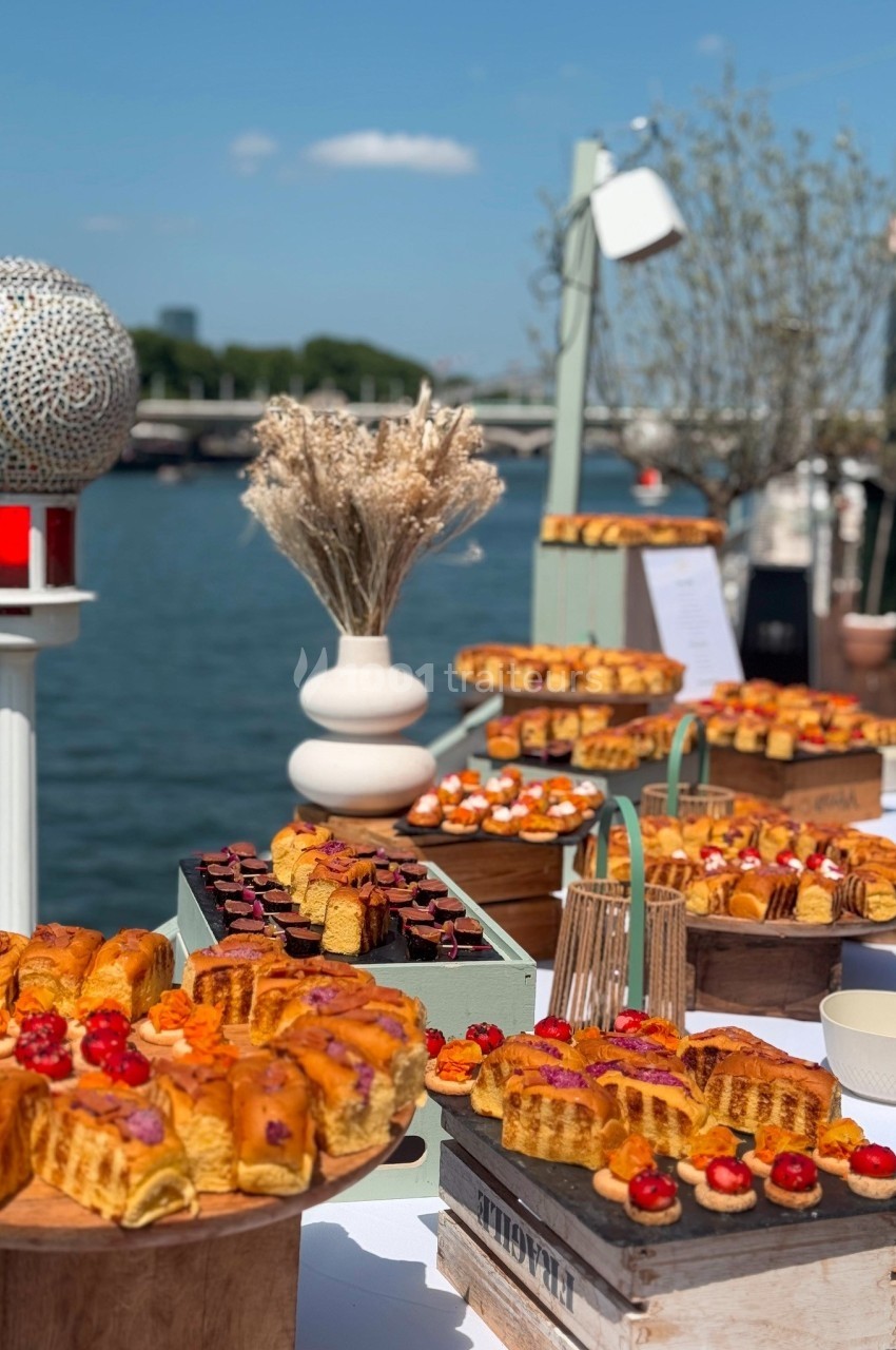Buffet en plein air avec viennoiseries, pâtisseries et décorations, installé près d'une rivière sous un ciel dégagé.