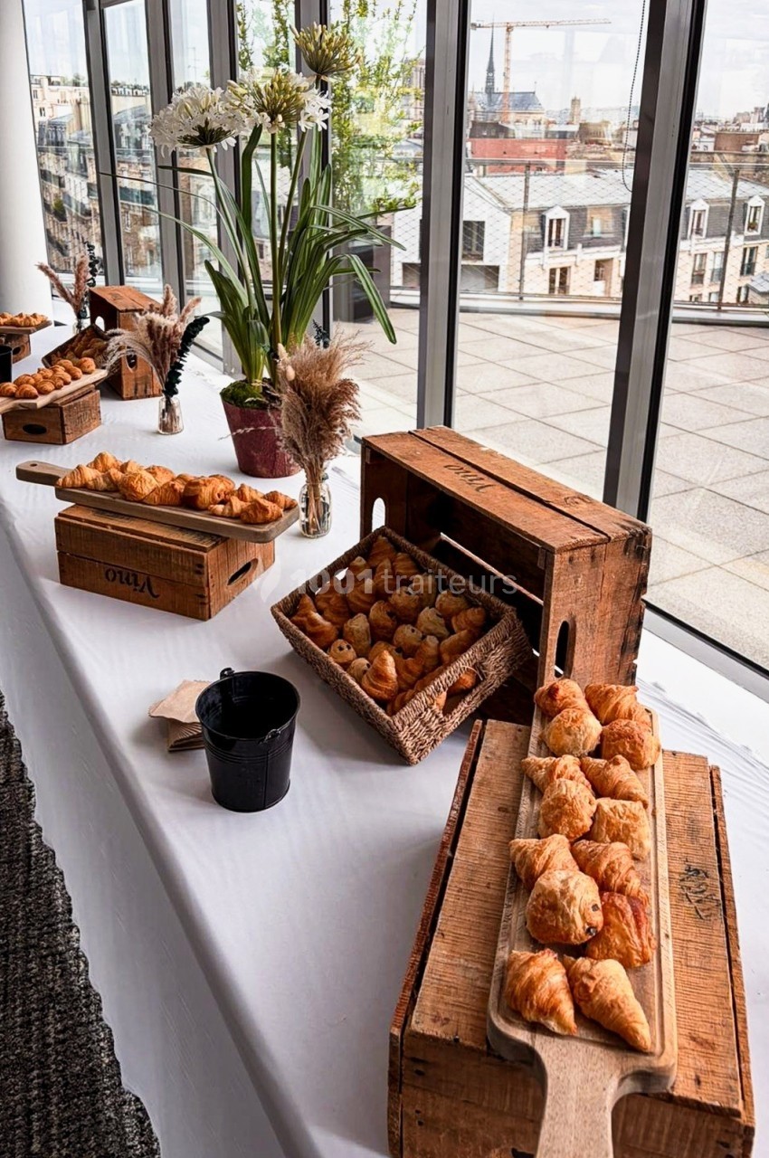 Table dressée avec des viennoiseries variées sur des plateaux en bois, près de grandes baies vitrées offrant une vue urbaine.