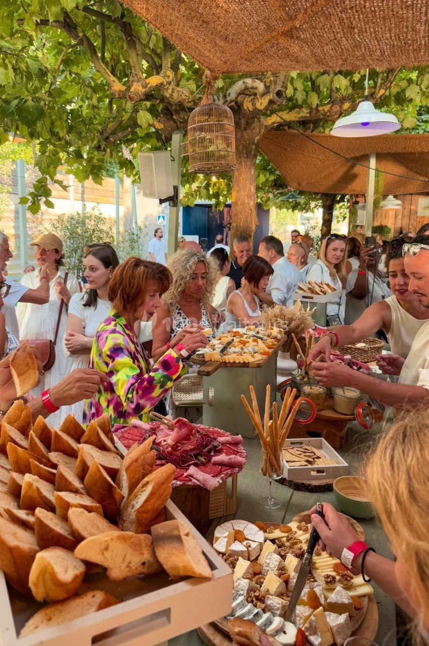 Buffet en plein air avec des invités se servant parmi des plats variés, sous une pergola ombragée entourée de verdure.