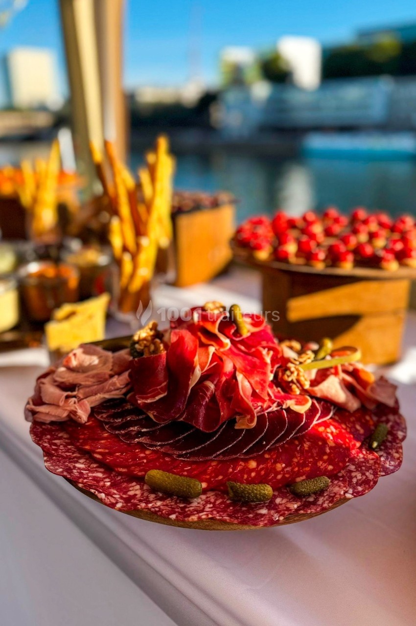 Plateau de charcuterie avec tranches de saucisson, jambon, cornichons et noix, présenté sur une table en extérieur.