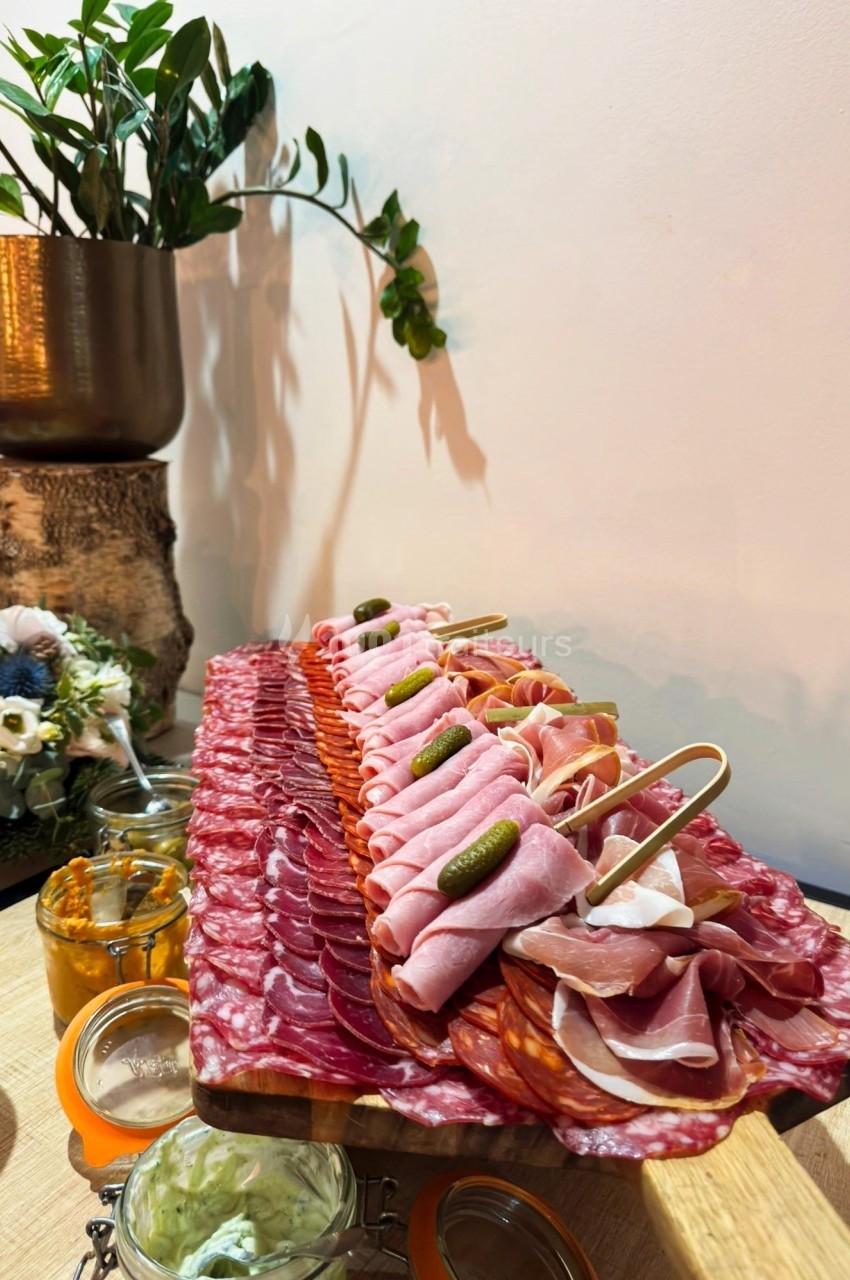 Plateau de charcuterie variée avec cornichons, présenté sur une table en bois, décorée de fleurs et plantes.