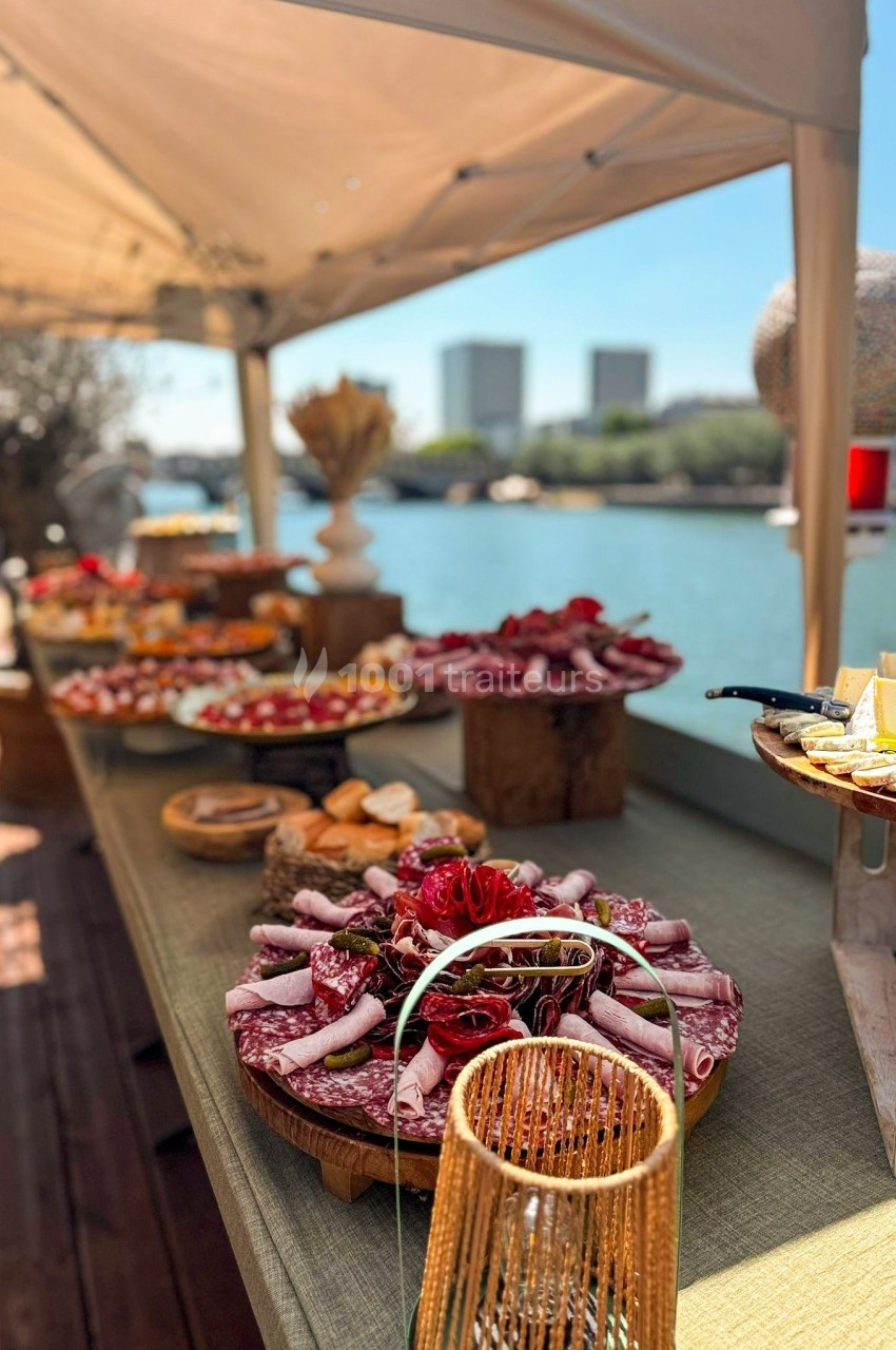 Table garnie de plats variés, incluant charcuterie et fromages, sous une tente près d'un plan d'eau urbain.