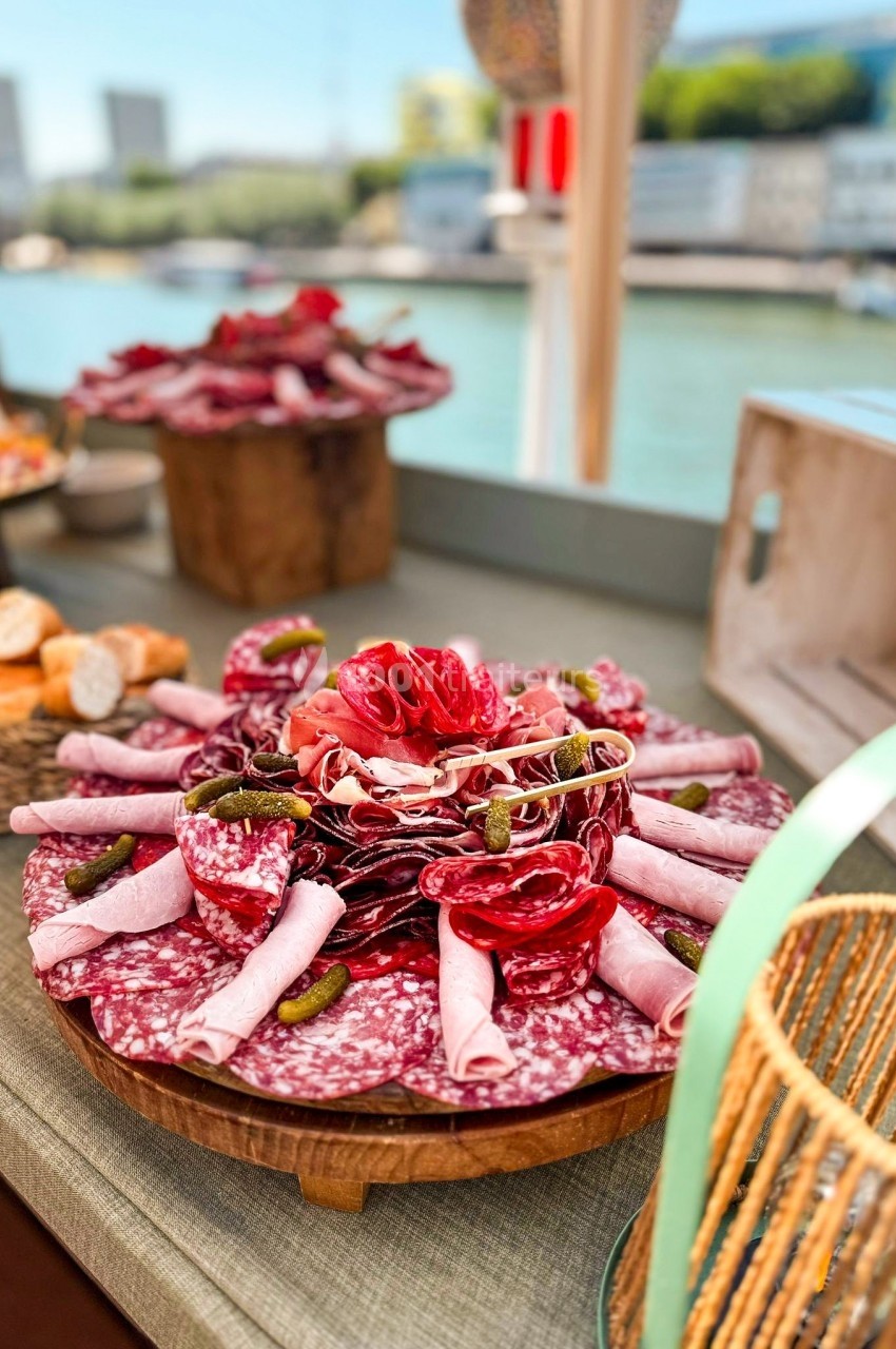 Plateau de charcuterie variée avec saucissons, jambons roulés et cornichons, présenté sur une table près d'une rivière.