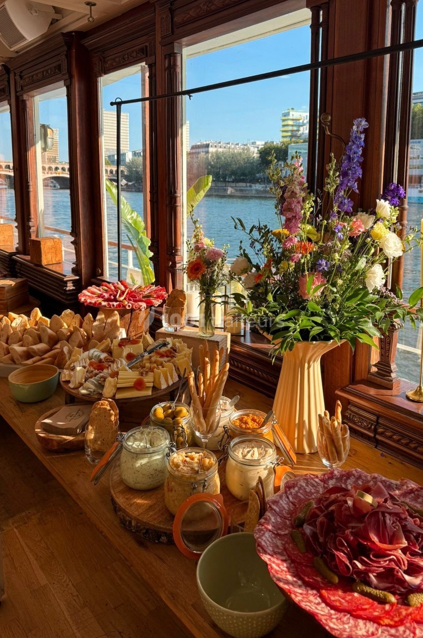 Buffet composé de charcuteries, pains, dips et fleurs, installé dans une salle lumineuse avec vue sur une rivière.