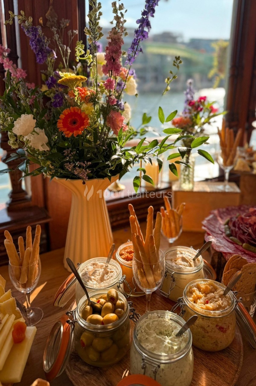 Plateau apéritif avec dips, olives, gressins et fleurs en arrière-plan près d'une fenêtre donnant sur un paysage ensoleillé.