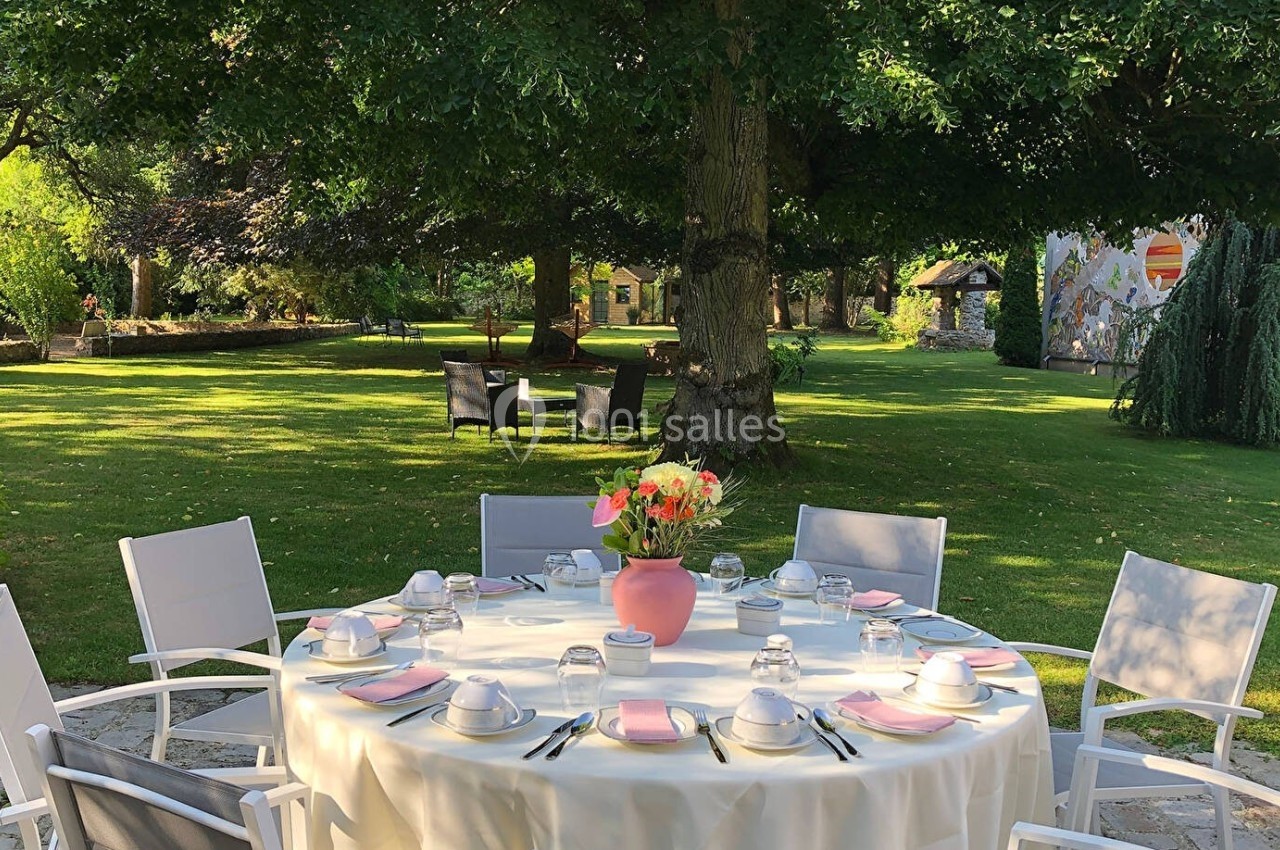 Table dressée avec vaisselle blanche et nappe beige dans un jardin verdoyant sous des arbres ombragés.