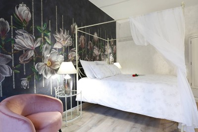 Lit à baldaquin blanc avec drapé, fauteuil rose et mur décoré de motifs floraux dans une chambre lumineuse.