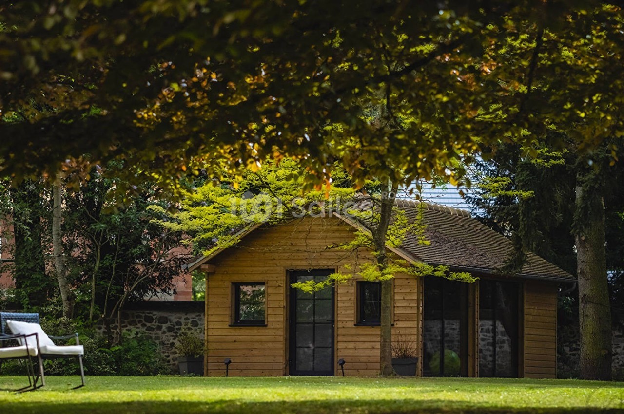 Abri de jardin en bois entouré de verdure, avec des arbres et une chaise blanche sur la pelouse au premier plan.