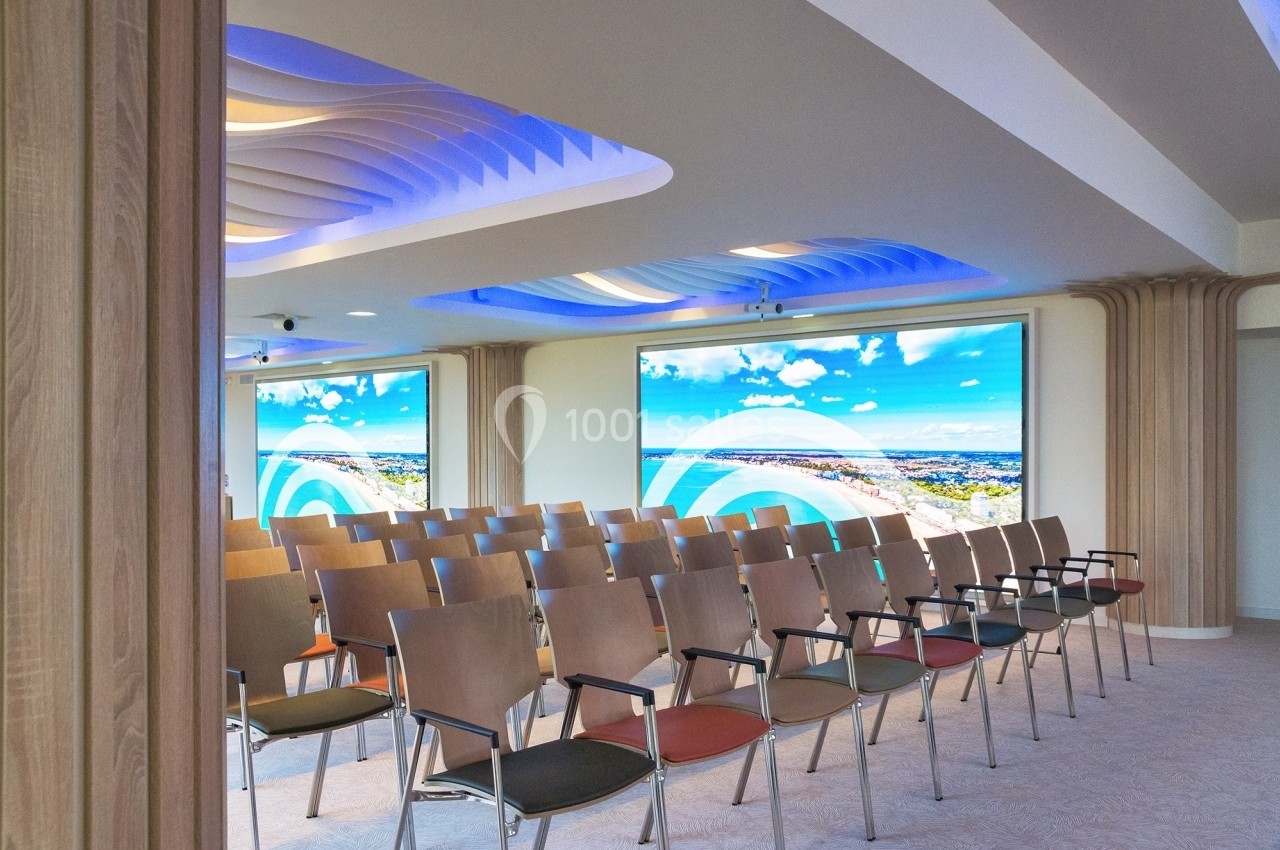 Salle de conférence moderne avec rangées de chaises en bois, écrans lumineux et plafond aux formes ondulées.