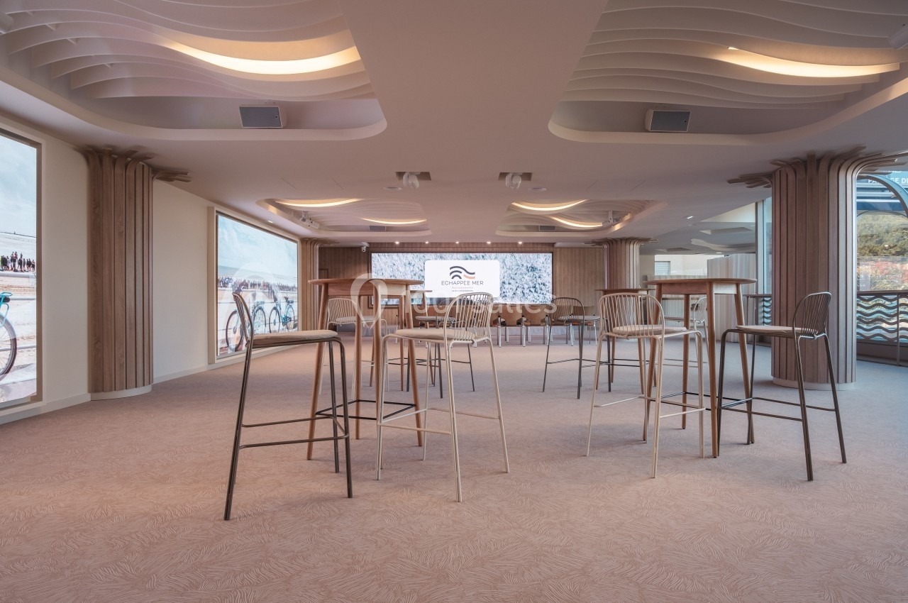 Salle de réunion lumineuse avec tables hautes, chaises et écran de présentation, décorée de panneaux muraux ondulés.