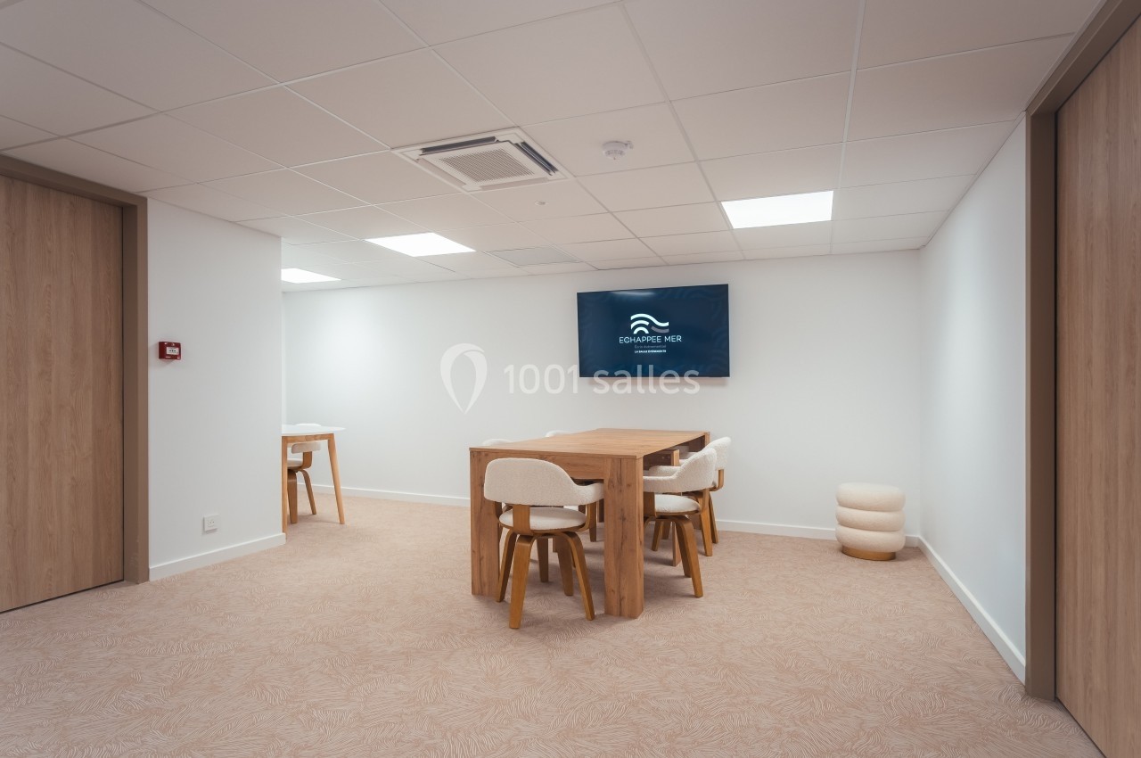 Salle de réunion lumineuse avec table en bois, chaises rembourrées et écran mural affichant un logo.