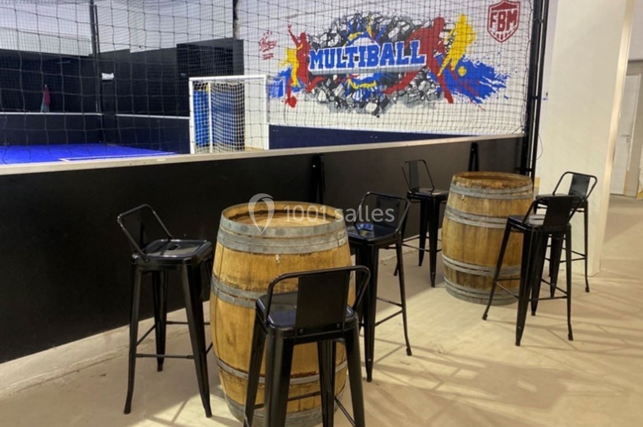 Tables en tonneaux avec chaises noires devant un terrain de sport intérieur entouré de filets.
