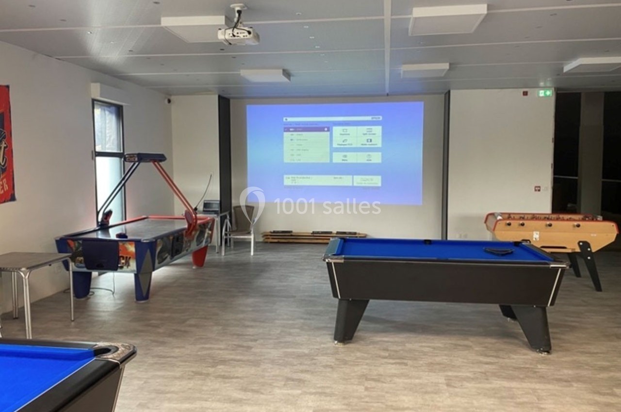 Salle de loisirs avec billards, baby-foot, jeu d'arcade et écran de projection allumé.