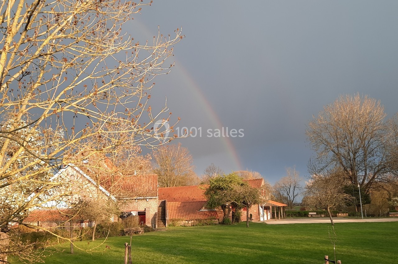 Location salle Linkebeek (Brabant flamand) - Ferme Holleken Hoeve #1 Un arc-en-ciel traverse un ciel nuageux au-dessus d'une maison en briques entourée d'arbres et d'une pelouse verte.