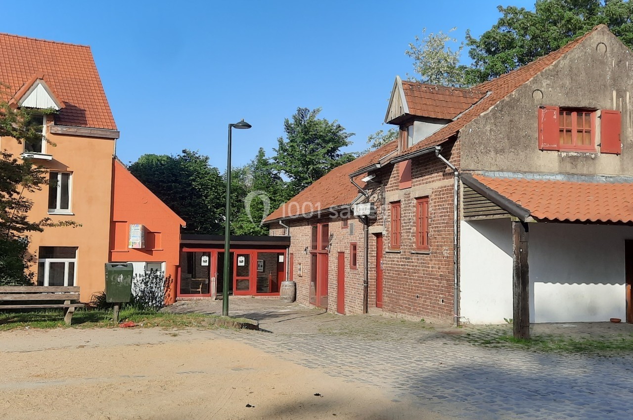 Location salle Linkebeek (Brabant flamand) - Ferme Holleken Hoeve #2 Bâtiments en briques rouges avec toits en tuiles, entourés d'arbres, sur une place pavée et ensoleillée.