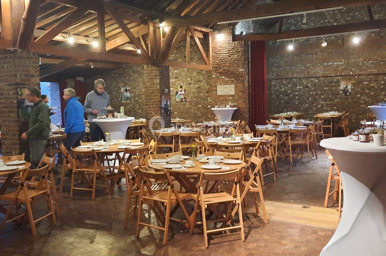 Location salle Linkebeek (Brabant flamand) - Ferme Holleken Hoeve #3 Salle en briques avec des tables rondes dressées pour un repas, quelques personnes en arrière-plan.