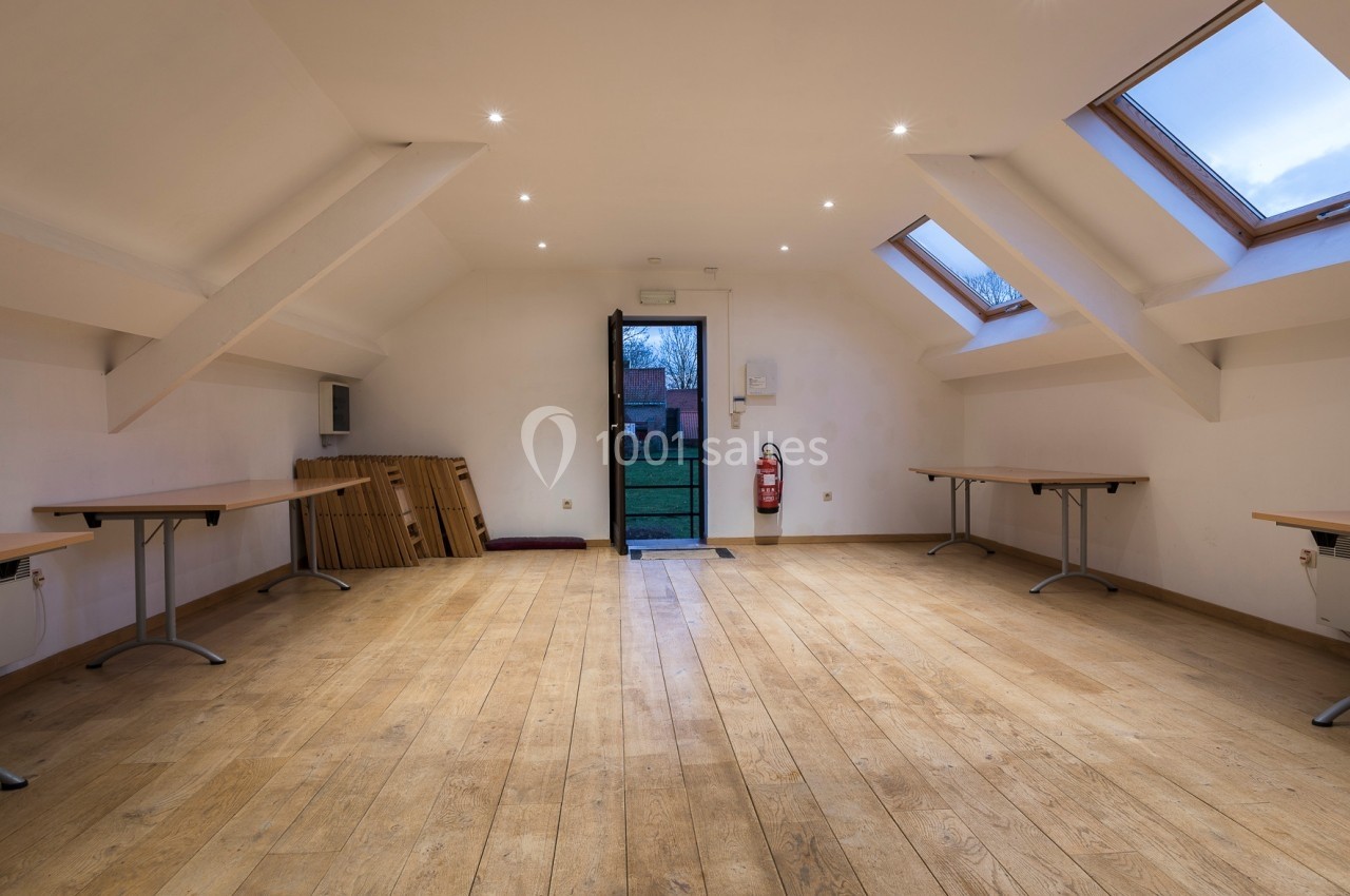 Location salle Linkebeek (Brabant flamand) - Ferme Holleken Hoeve #11 Salle vide avec parquet en bois, tables pliées contre un mur, fenêtres de toit et porte ouverte donnant sur l'extérieur.
