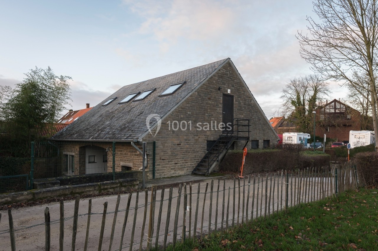 Location salle Linkebeek (Brabant flamand) - Ferme Holleken Hoeve #4 Bâtiment en pierre avec toit incliné, escaliers extérieurs et clôture en bois, entouré de végétation et de maisons.