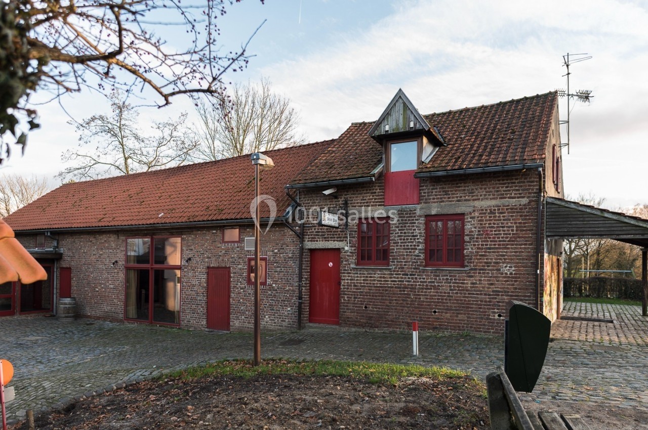 Location salle Linkebeek (Brabant flamand) - Ferme Holleken Hoeve #5 Bâtiment en briques rouges avec toit en tuiles, fenêtres rouges et petite tour, entouré de pavés et d'arbres.