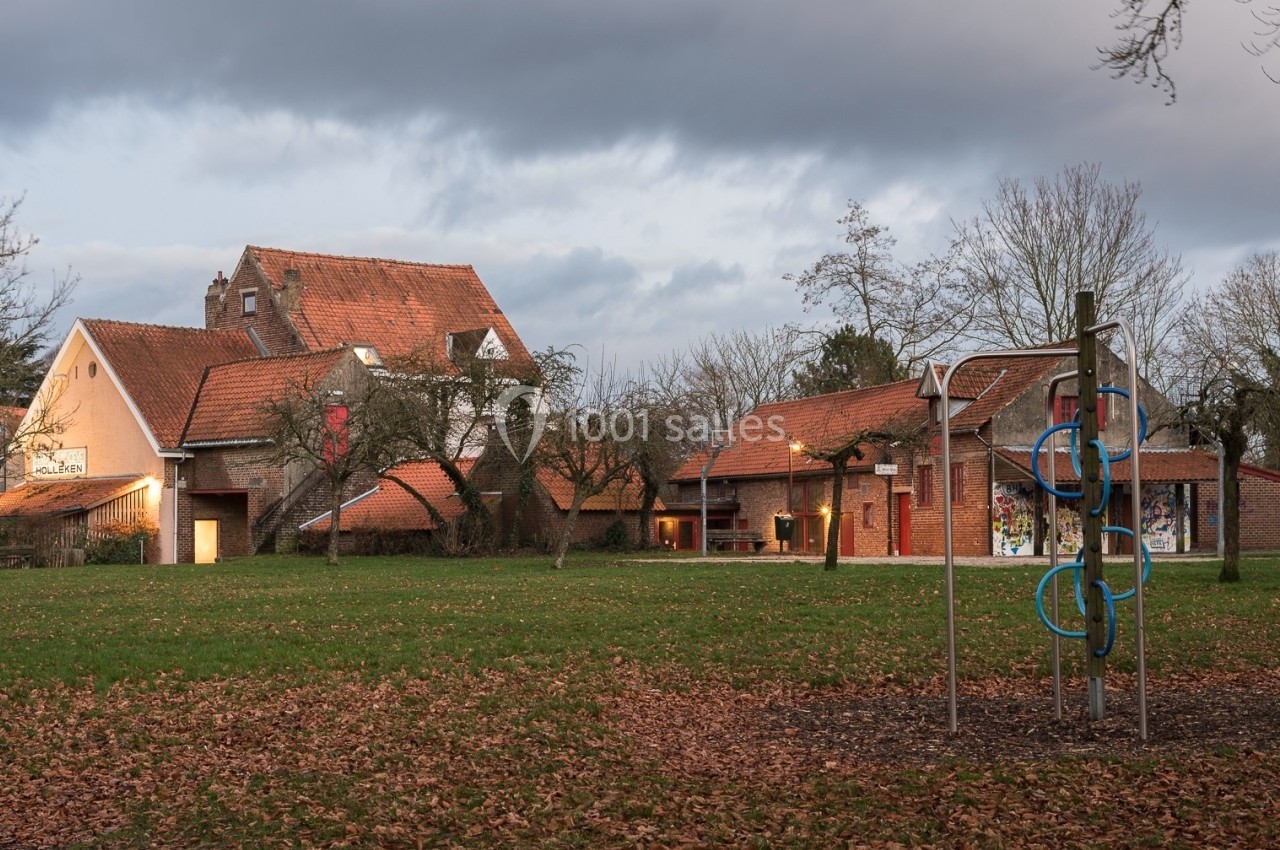 Location salle Linkebeek (Brabant flamand) - Ferme Holleken Hoeve #6 Bâtiments en briques rouges avec toits en tuiles entourés d'arbres, aire de jeux au premier plan sur une pelouse.